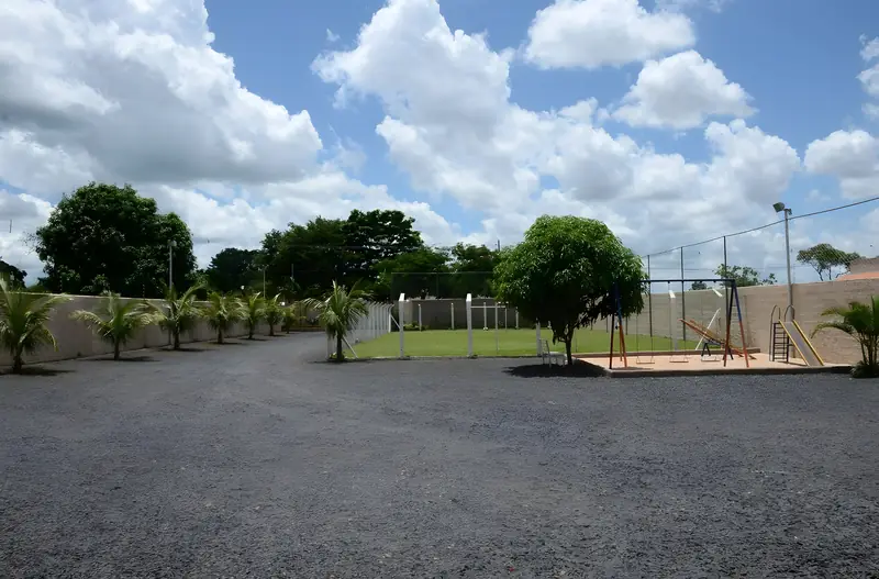 Estacionamento amplo de brita, com gramado, árvores e o parquinho ao fundo