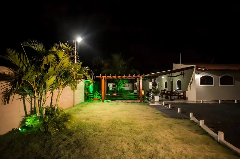 Jardim iluminado à noite, com palmeiras, pérgola e a casa ao fundo