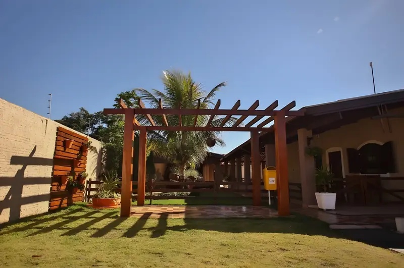 Pérgola de madeira sobre o gramado, com a varanda da casa ao fundo