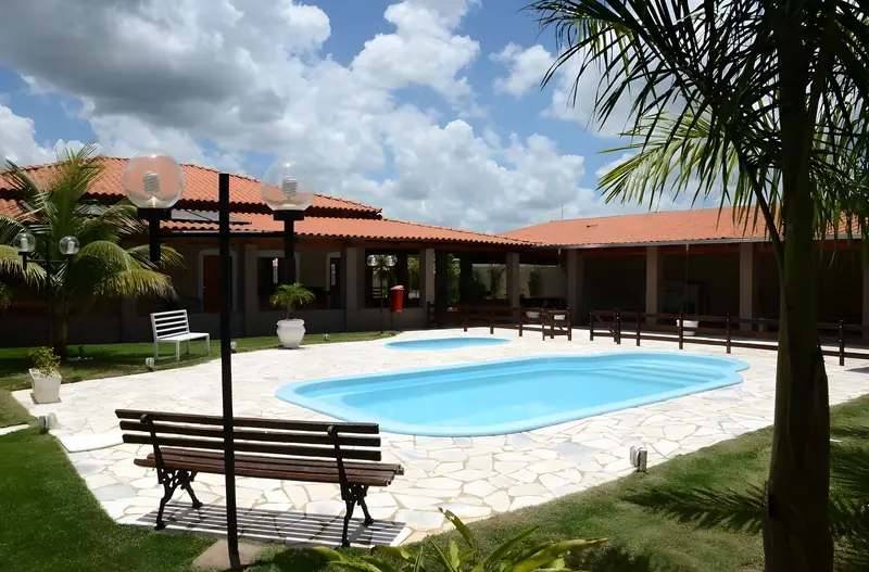 Piscina da Chácara Império com banco de madeira, palmeira e a casa ao fundo, em dia de céu azul